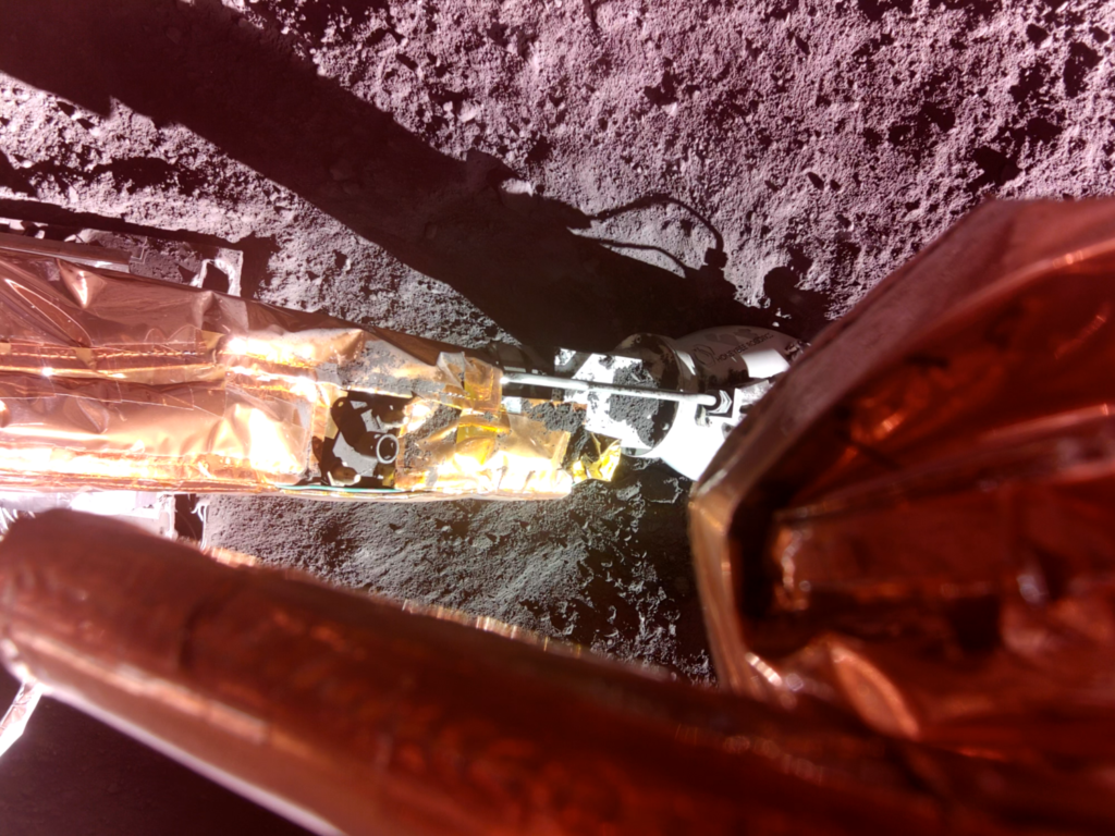 A robotic arm extends onto a gray, rocky lunar surface. At the end of the arm is the Lunar PlanetVac, a small, cylindrical device. The scene is illuminated, showing details of the lunar surface and the lander's components. Image credit: Firefly Aerospace, via fireflyspace.com