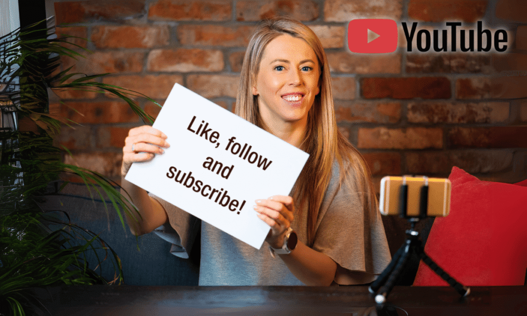 Female YouTube creator in home studio setting holding "Like, follow and subscribe!" sign with smartphone on tripod and brick wall background, illustrating creator engagement strategies that helped build YouTube's 2.7 billion user community.